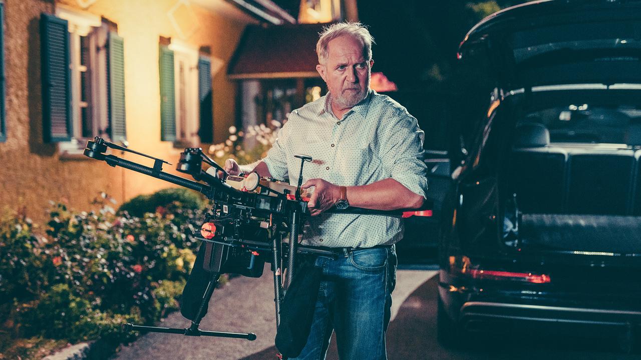 © ORF/Steirerwahn/Stefan Robitsch Harald Krassnitzer hält eine große Drohne, neben einem Auto bei Nacht.
