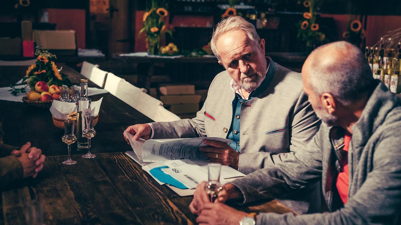 © ORF/Steirerwahn/Stefan Robitsch Harald Krassnitzer blättert in Unterlagen und blickt Simon Hatzl an, während sie an einem rustikalen Holztisch mit Schnapsgläsern sitzen.