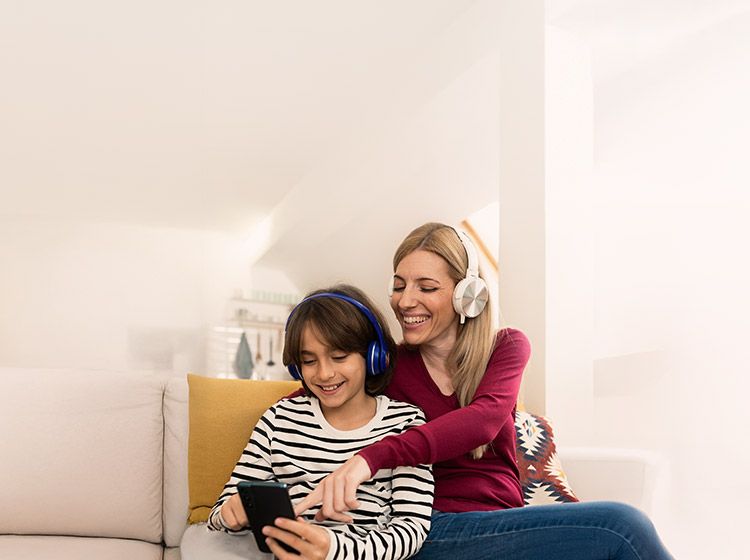 Mutter und Sohn sitzen auf Sofa; nutzen Smartphone mit Kopfhörern