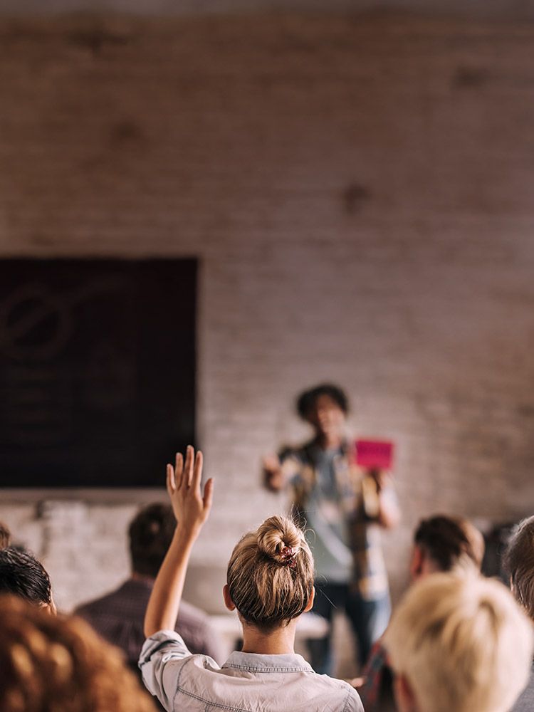 Warme Aufnahme eines Business Vortrags. Eine Person steht auf der Bühne und gestikuliert. Viele Anwesende die interessiert zuhören - man sieht sie nur von hinten. Eine Frau mit blonden Haaren zeigt auf.