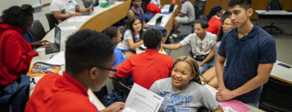 A diverse group of students in a classroom engaged in discussion and collaborative work. Some are using laptops, and others are reviewing documents. The setting is casual and interactive, suggesting a study session or group project.