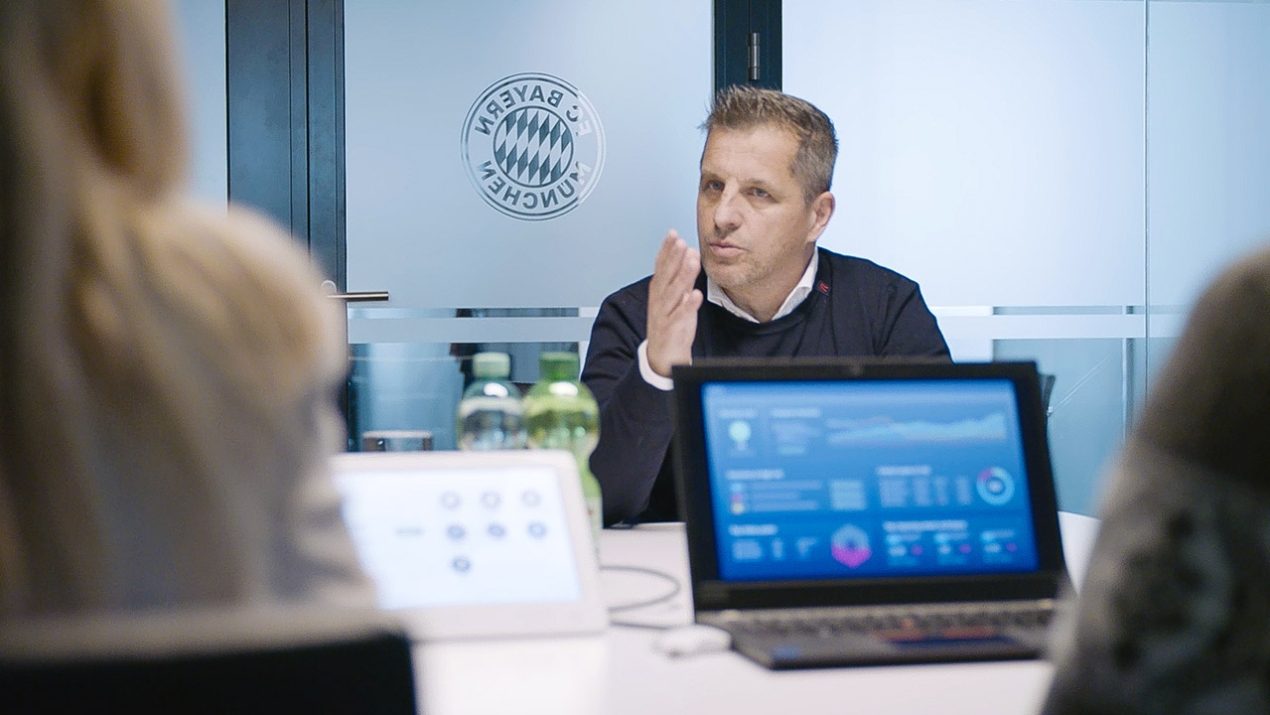 The picture shows a meeting. A door with the FC Bayern Munich logo can be seen in the background. A white, middle-aged man is sitting in front of the door. In the foreground, you can see the back of a woman with long blonde hair and, to the right, the shoulder of another person. It can be seen that the people are talking, as there is a laptop on the table showing diagrams.